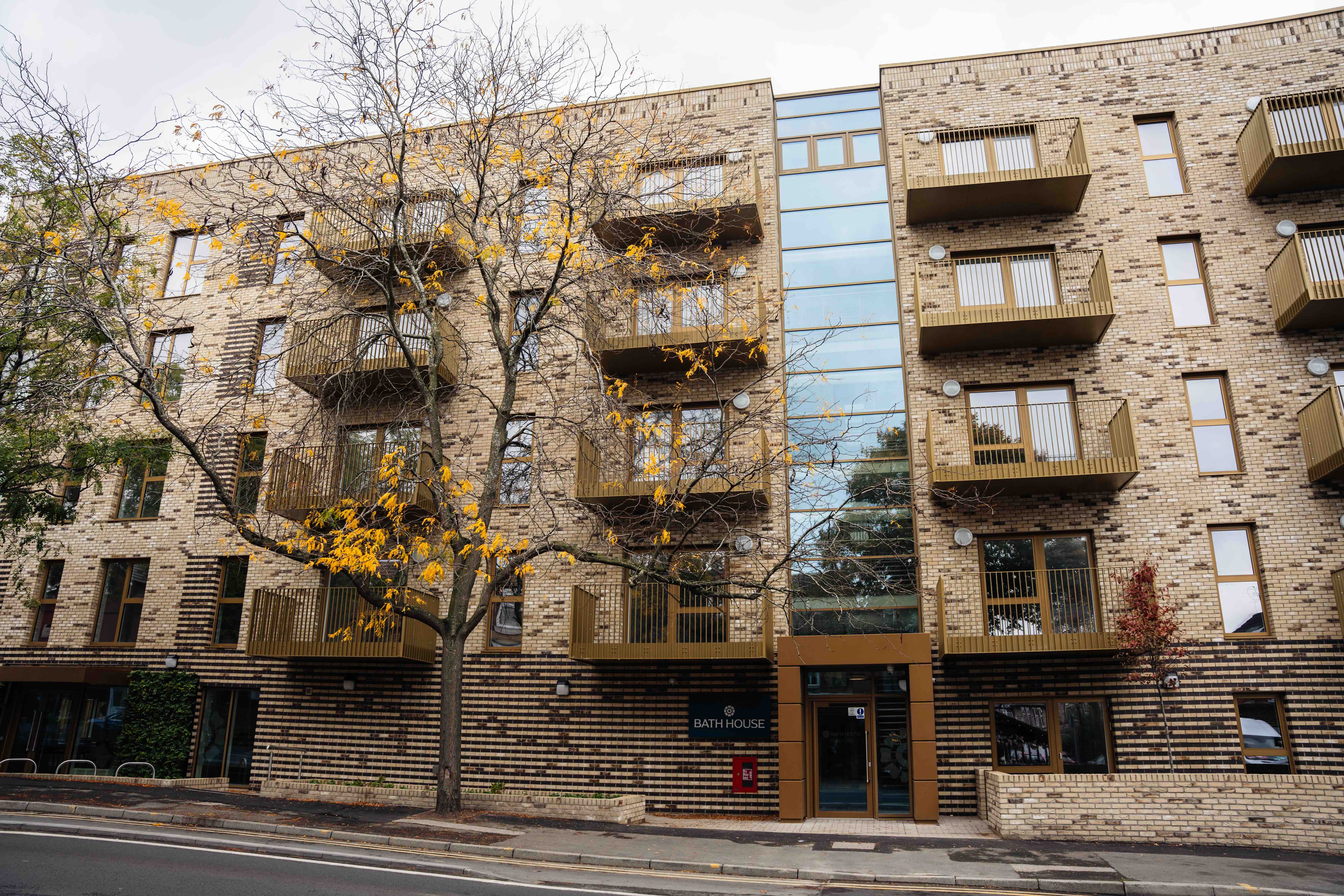 Bath House external view facing the road with balconies and a large tree with yellowing leaves